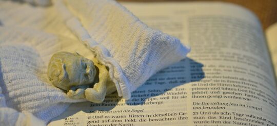kleine Figur Kind gewickelt, liegt auf einer aufgeschlagenen Bibelseite. Foto: pixabay.com, pfarrbriefservice.de